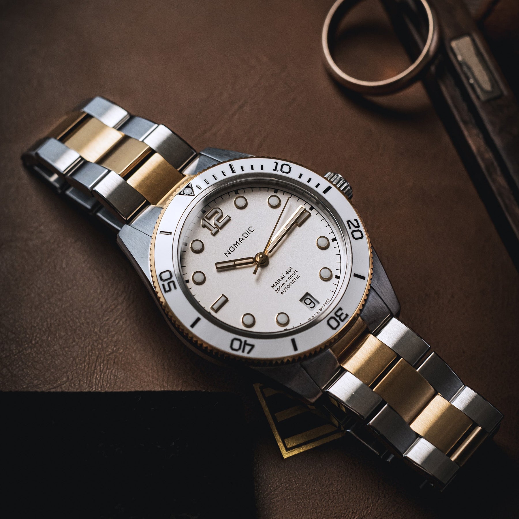 A two-tone stainless steel and gold wristwatch with a white dial, resting on a dark brown leather surface next to a gold ring.