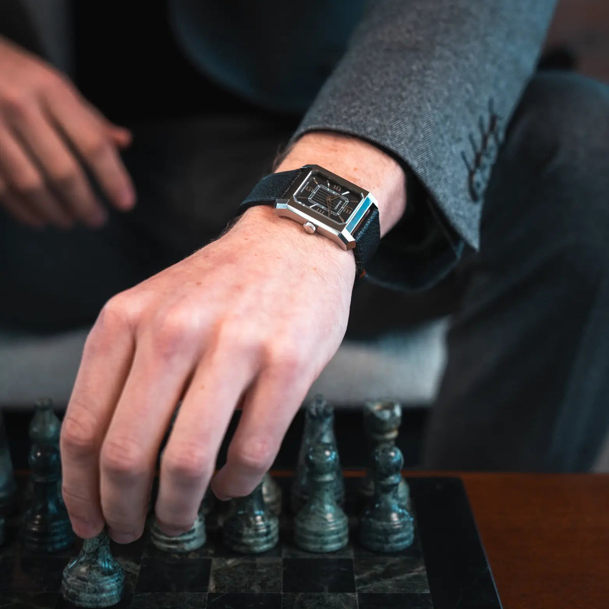 A close-up lifestyle shot of a man's hand resting on a chessboard while wearing a modern rectangular smartwatch with a black strap and silver-toned case.
