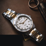 A two-tone stainless steel and gold wristwatch with a white dial, resting on a dark brown leather surface next to a gold ring.