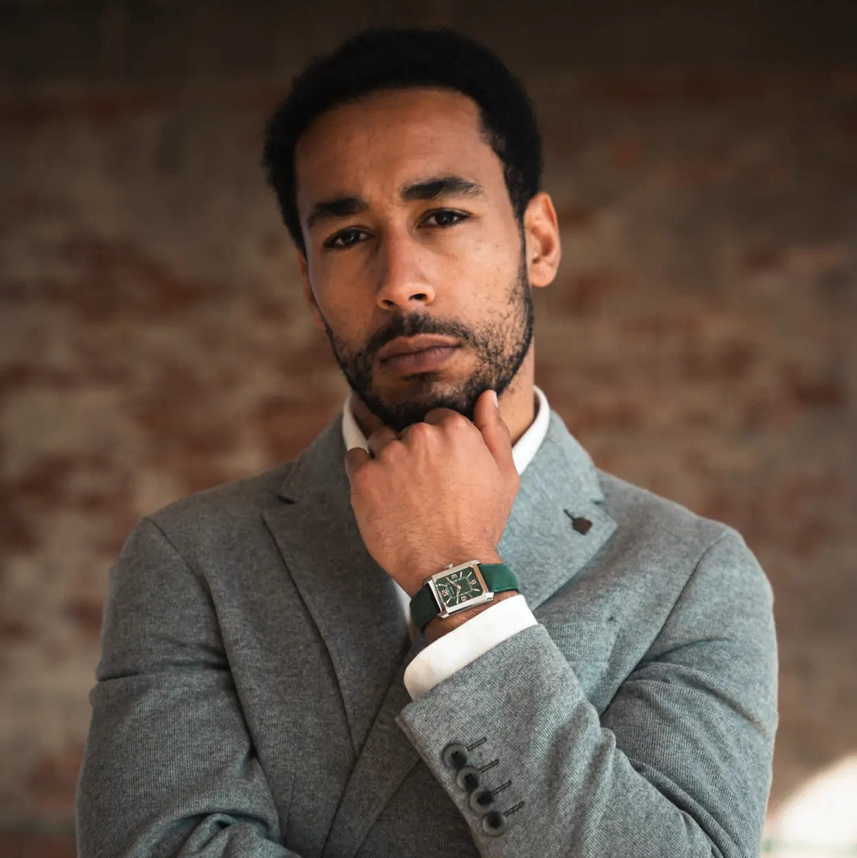 A man wearing a textured gray blazer over a white collared shirt, with a green leather strap watch visible on his wrist, posing against a rustic brick wall background.